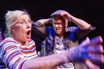 Silmälasipäinen ja raitapaitainen nainen näyttää järkyttyneeltä suu auki, kun taas hänen takanaan oleva mies pitää hattua jännittyneenä ja huolestuneena. Molemmat ovat dramaattisen näyttämövalaistuksen alla.