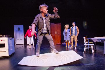 Mies tasapainoilee näyttämöllä jättimäisen avoimen kirjan päällä, kun taas kolme muuta ihmistä seisoo taustalla keittiökalusteiden ja rekvisiitan vieressä dramaattisessa valaistuksessa teatteriympäristössä.