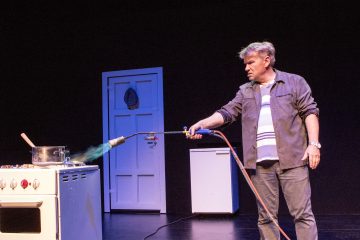 Rennosti pukeutunut mies käyttää puhalluslamppua liedellä olevaan kattilaan, jonka sisällä on puulusikka. Kohtaus näyttää olevan lavalla, jonka taustalla on ovi ja pieni jääkaappi.