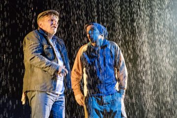 Kaksi takki- ja hattupäistä miestä seisoo tiiviisti vierekkäin rankkasateessa lavavalojen valaisemina, ja vettä valuu pimeässä näkyvästi heidän ympärilleen.