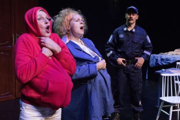 Kaksi naista, toinen punaisessa hupparissa ja toinen sinisessä takissa, jolla on tekoraskausvatsa, seisovat lavalla silmät suurina ja suu auki, kun poliisin univormuun pukeutunut mies katsoo heitä. Kohtaus vaikuttaa dramaattiselta tai koomiselta.