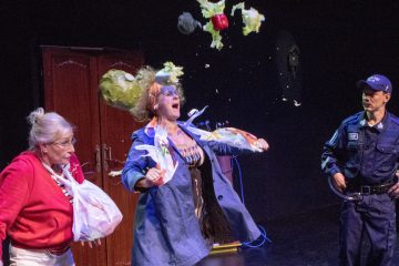 Lavalla on kolme ihmistä: yllättynyt vanhempi nainen punaisessa takissa, jolla on kädessään kassit, sinipukuinen henkilö, jonka pään yli lentää ruokaa ja salaattia, ja univormuun pukeutunut poliisi, joka seuraa tapahtumia.