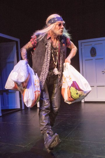 Henkilö, jolla on pitkät valkoiset hiukset, päässään otsanauha, aurinkolasit, liivit ja nahkahousut, kantaa kahta suurta muovista ostoskassia lavalla, jonka taustalla on valkoinen ovi ja rekvisiittaa.