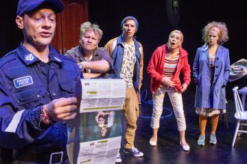 Poliisi lukee sanomalehteä ääneen neljälle yllättyneelle ja huolestuneelle aikuiselle, jotka seisovat hänen takanaan näyttämöllä, mikä viittaa dramaattiseen tai komedialliseen teatterikohtaukseen.