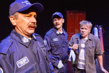 Sisällä seisoo kolme miestä; kahdella on sininen poliisiasu ja hattu, ja yhdellä on harmaa paita ja takki. Tausta on tumma, ja heidän takanaan näkyy puinen ovi ja sylinterit. Kohtaus vaikuttaa jännittyneeltä tai vakavalta.