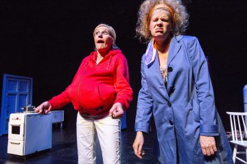 Kaksi näyttelijää näyttämöllä tekee liioiteltuja ilmeitä; toisella on punainen huppari ja valkoiset housut, toisella sininen takki ja sotkuiset hiukset. Taustalla näkyy liesi, tuoli ja ovi, jotka viittaavat keittiöön.