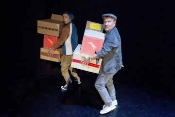 Kaksi rennosti pukeutunutta, takkeihin ja hattuihin pukeutunutta miestä kantaa suuria pahvilaatikoita, joissa on punaiset dinosauruslogot, dramaattisessa valaistuksessa tummalla taustalla ja katsoo olkapäitään vasten.