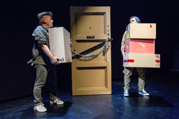 Kaksi ihmistä seisoo lavalla pitelemässä suuria laatikoita, ja heitä vastakkain on kahlehdittu asunnon ovi. Ympäristö on hämärästi valaistu, mikä viittaa teatteriesitykseen tai dramaattiseen kohtaukseen, johon liittyy liikkumista tai yhteenottoa.