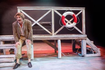 Mies, jolla on sotkuinen tukka, viikset ja rennot vanhanaikaiset vaatteet, seisoo puisella laiturilla, jossa on pelastusrengas. Hän nojaa eteenpäin ja näyttää puhuvan tai näyttelevän teatteriesityksessä.