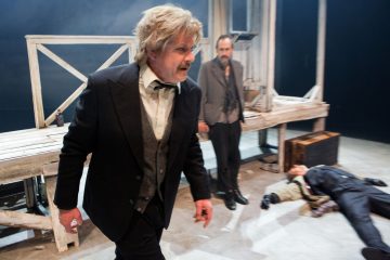 Pukumies kävelee vihaisena eteenpäin kuistia muistuttavalla lavasteella, kun toinen mies seisoo taustalla ja kolmas henkilö makaa liikkumattomana lattialla. Kohtaus on dramaattinen ja jännittynyt.