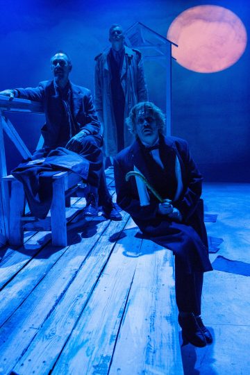 Kolme tummiin takkeihin pukeutunutta miestä on lavasteessa, jossa on puisia lankkuja ja penkkejä, joita valaisee sininen valo. Yksi mies istuu keppi kädessään, toinen istuu penkillä, ja kolmas seisoo heidän takanaan, ja taustalla on suuri oranssi kuu.