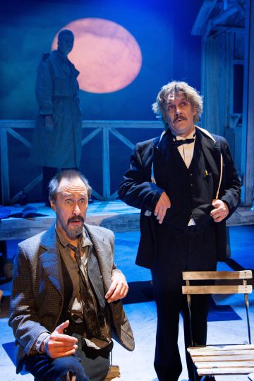 Kaksi vanhanaikaiseen asuun pukeutunutta miestä, toinen istuen ja toinen seisten, esiintyy teatterilavalla. Taustalla seisoo kolmas juoksutakkiin pukeutunut hahmo, jonka taakse on heijastettu suuri vaaleanpunainen kuu.
