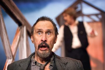 Mies, jolla on suuret siniset silmät ja yllättynyt ilme, seisoo terävänä, kun taas toinen liiviin pukeutunut henkilö on sumeana taustalla dramaattisessa lavastuksessa.