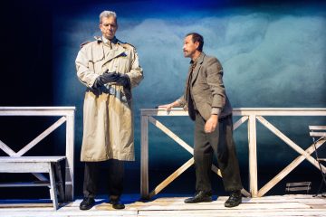 Kaksi miestä seisoo puisella lavalla, jossa on sininen tausta; toisella on trenssitakki ja hanskat, ja hän näyttää vakavalta, kun taas toinen nojaa kaiteeseen, on pukeutunut rennosti ja vaikuttaa jännittyneeltä.