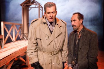 Lavalla seisoo kaksi miestä; toisella on beige trenssitakki ja hanskat, ja hän katsoo alaspäin, kun taas toinen, kuluneessa takissa, katsoo häntä. Puurakenne ja sumuinen tausta viittaavat dramaattiseen kohtaukseen.