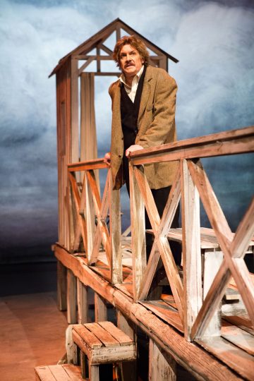 Ruskeaan takkiin ja mustaan liiviin pukeutunut mies, jolla on aaltoilevat hiukset ja viikset, seisoo lavalla puisen sillan kaltaisen rakenteen päällä, ja hänen takanaan on pilvinen taivas.