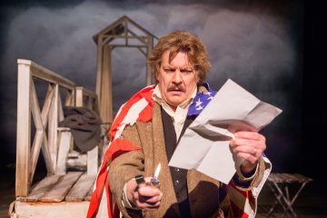 Amerikan lippuun verhoutunut mies pitää toisessa kädessään sytytintä ja toisessa rypistynyttä paperia seisoen hämärästi valaistulla näyttämöllä olevan puurakenteen edessä.