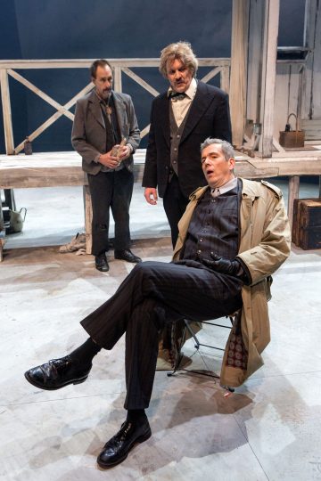 Lavalla on kolme aikakauden pukuihin pukeutunutta miestä. Yksi istuu tuolilla pitkä takki yllään, ja kaksi muuta seisovat hänen takanaan. Lavastuksessa on maalaismaisia puuelementtejä ja matkatavaroita, jotka tuovat mieleen 1900-luvun alun näyttämön.