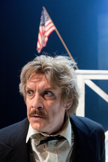 Mies, jolla on sotkuiset vaaleat hiukset ja viikset, puku ja rusetti, seisoo valkoisen puurakennelman edessä, jonka taustalla on Amerikan lippu. Hän katsoo sivulle vakavalla ilmeellä.