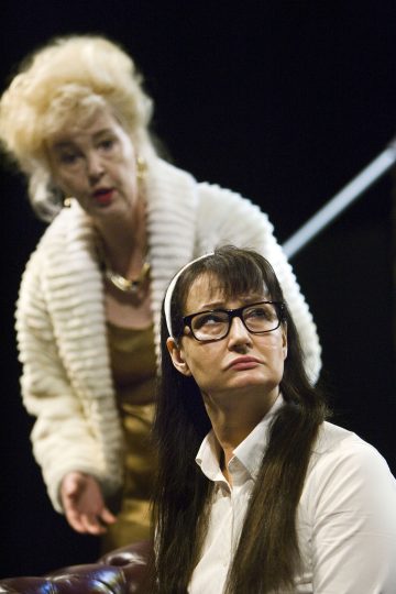 Tummahiuksinen, silmälasipäinen, valkoiseen paitaan pukeutunut nainen istuu katsoen ylöspäin vakavalla ilmeellä, ja hänen takanaan seisoo vaalea, valkoiseen turkkiin pukeutunut nainen, joka puhuu vilkkaasti.