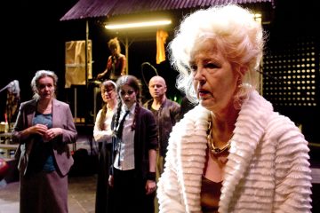 Etualalla seisoo vaaleahiuksinen, valkoiseen turkistakkiin pukeutunut nainen, joka näyttää vakavalta, ja hänen takanaan seisoo neljä dramaattisiin pukuihin pukeutunutta ihmistä hämärästi valaistussa lavastuksessa.