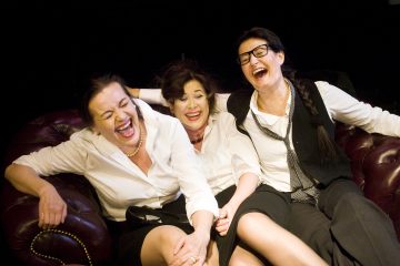 Kolme liikemieheksi pukeutunutta naista istuu lähekkäin ruskealla nahkasohvalla ja nauraa iloisesti. Tausta on tumma, mikä korostaa heidän iloisia ilmeitään ja vilkasta vuorovaikutusta.