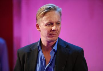 Lyhyet vaaleat hiukset omaava mies, jolla on tumma bleiseri ja sininen paita, seisoo vaaleanpunaisen ja violetin taustan edessä ja katsoo hieman sivulle neutraali ilme kasvoillaan.