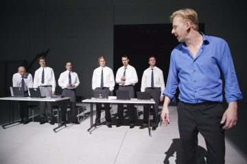 Kuusi valkoisiin paitoihin ja solmioihin pukeutunutta miestä seisoo työpöytien takana kannettavien tietokoneiden kanssa, kun taas sinipukuinen mies seisoo etualalla ja katsoo heitä kohti kirkkaasti valaistussa, minimalistisessa ympäristössä.