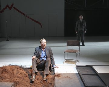 Pukumies istuu lavalla likaisen kuopan reunalla, toinen jalka sisällä ja toinen ulkona. Lähellä on tyhjä tuoli, ja toinen mies seisoo taustalla. Lavastus on minimaalinen, ja seinällä on punainen siksak-viiva.