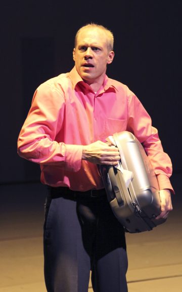 Kirkkaan vaaleanpunaiseen paitaan pukeutunut mies, joka seisoo valonheittimen alla muuten pimeässä huoneessa, pitää kädessään harmaata radiopuhelinta ja joitakin papereita ja näyttää huolestuneelta tai yllättyneeltä.