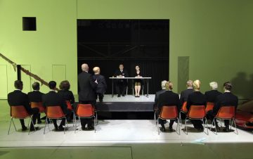 Joukko mustiin pukuihin pukeutuneita ihmisiä istuu oransseilla tuoleilla näyttämölle päin, jossa kaksi ihmistä istuu pöydän ääressä. Paikka on sisätiloissa, joissa on minimaalinen, moderni sisustus ja kova valaistus.