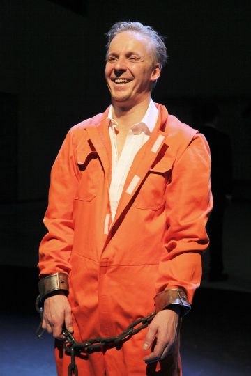 Hymyilevä mies oranssissa vankilahaalarissa ja valkoisessa paidassa seisoo ranteet kahlittuina ja kädet yhteen kahlittuina tumman taustan edessä.