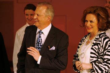 Tummassa puvussa ja kuviollisessa solmiossa oleva mies hymyilee seisoessaan seepra-raidalliseen takkiin, helmiin ja punaiseen huulipunaan pukeutuneen naisen vieressä. He näyttävät olevan juhlatilaisuudessa, ja taustalla hymyilee muita.