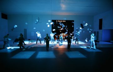 Ryhmä ihmisiä hämärässä, sinisessä valossa kylpevällä näyttämöllä, ja heidän yläpuolellaan lentää dramaattisesti ilmassa lukuisia paperiarkkeja.