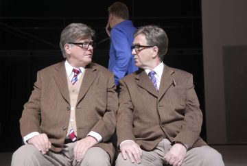 Kaksi vanhempaa miestä, joilla on harmaat hiukset, silmälasit ja ruskeat puvut, istuvat vierekkäin ja katsovat toisiaan. Molemmilla on raidallinen solmio ja beiget housut. Kolmas sinipukuinen henkilö seisoo taustalla, kasvot poispäin.