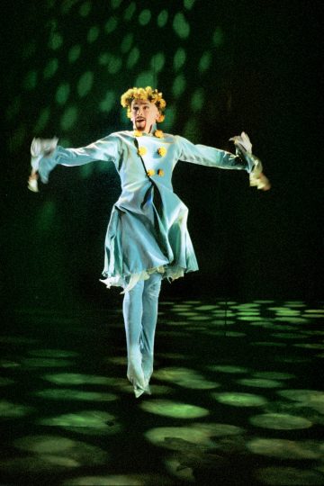 Vaaleansiniseen mekkoon, housuihin ja kukkakruunuun pukeutunut henkilö tanssii tarmokkaasti lavalla, jossa vihreät valonheittimet luovat pyöreitä kuvioita lattiaan ja taustalle.