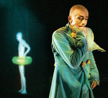 Lavalla seisoo kalju esiintyjä, jolla on sininen, kukkakoristeinen puku, toinen käsi kasvojensa lähellä. Taustalla näkyy hämärä, hehkuva ihmishahmo hämärästi tummalla taustalla.