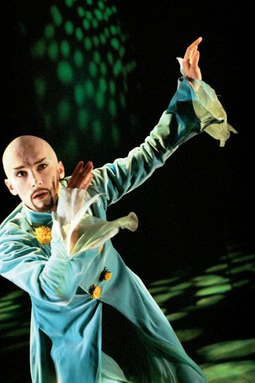 Kaljupäinen ja vuohenpukin omaava esiintyjä, joka on pukeutunut virtaavaan turkoosiin pukuun, jossa on röyhelöiset hihat ja keltaisia kukkia, poseeraa dramaattisesti vihreiden näyttämövalojen alla.