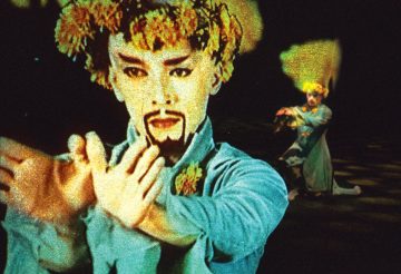 Hienostuneesti meikattu ja puvustettu esiintyjä, jolla on kukkia päässään, elehtii dramaattisesti, kun taas toinen, samoin pukeutunut hahmo poseeraa taustalla pimeällä näyttämöllä.