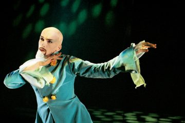 Dramaattiseen meikkiin ja keltaisilla korostuksilla varustettuun tiilenpunaiseen pukuun pukeutunut esiintyjä poseeraa näyttämöllä tunnelmallisessa, vihreän sävyisessä valaistuksessa.