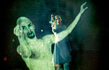 Dramaturgisesti maskeerattu ja puvustettu esiintyjä seisoo toinen käsi kohotettuna, kun hänen vieressään näkyy suuri, aavemainen, puoliksi läpinäkyvä projektio hänen kuvastaan tummaa, eteeristä taustaa vasten.