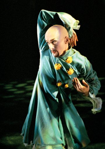 Esiintyjä, joka on pukeutunut virtaavaan sinivihreään kaapuun ja keltaisiin kukkien kaltaisiin koristeisiin, poseeraa dramaattisesti lavalla, toinen käsi taivutettuna pään yläpuolelle ja ilme keskittyneenä tummaa taustaa vasten.