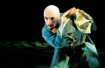 Keltaisilla kukkasilla ja läpinäkyvillä hihoilla koristeltuun tiilenpunaiseen pukuun pukeutunut esiintyjä poseeraa dramaattisesti lavalla, ja hänellä on ajeltu pää ja pukinparta tummaa taustaa vasten.