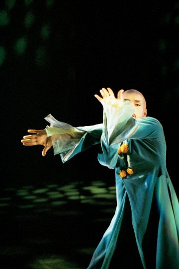 Henkilö, jolla on virtaava sininen puku, jossa on läpikuultavat hihat, poseeraa dramaattisesti ja peittää kasvonsa osittain käsillään tummaa, varjoista taustaa vasten.