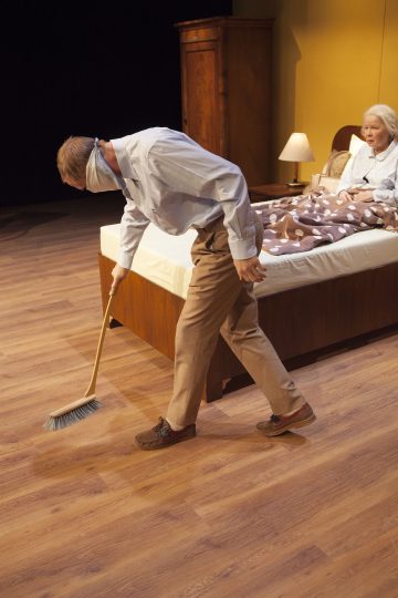 Silmät sidottu henkilö lakaisee lattiaa sängyn vieressä, kun vanhempi nainen istuu sängyssä ruskean peiton alla ja katsoo. Huoneessa on puiset huonekalut ja lämmin valaistus.