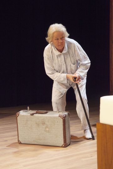 Vaaleaan pyjamaan pukeutunut iäkäs nainen seisoo lavalla, nojaa keppiin ja katsoo alas jalkojensa juurella olevaa vanhaa matkalaukkua. Tausta on tumma.