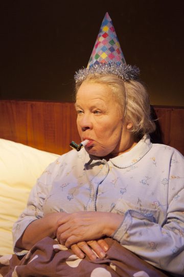 Pyjama-asuinen iäkäs nainen istuu sängyssä värikäs juhlahattu päässään, puhaltaa juhlatorveen ja hänen ilmeensä on neutraali.