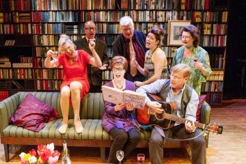 Kuusi ihmistä laulaa innokkaasti yhdessä viihtyisässä kirjahyllyjen reunustamassa huoneessa; yksi soittaa kitaraa, toiset pitävät juomia kädessään, ja kaksi istuu vihreällä sohvalla, kun muut seisovat takana, ja kaikki vaikuttavat iloisilta ja eloisilta.