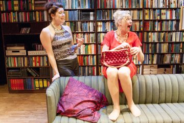 Kaksi naista on huoneessa, jossa on kirjahyllyjä. Toisella naisella, joka seisoo ja hymyilee, on hihaton toppi ja mustat housut. Toinen nainen, jolla on punainen mekko, istuu paljain jaloin vihreällä sohvalla, nauraa ja pitää kädessään punaista käsilaukkua.