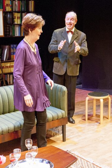 Lila toppapukuinen nainen seisoo vihreän sohvan vieressä ja katsoo alaspäin, kun taas pukuinen ja rusettisolmioinen mies elehtii vilkkaasti hänen takanaan. Viihtyisässä, hyvin valaistussa huoneessa näkyy kirjahyllyjä ja pieni pöytä.